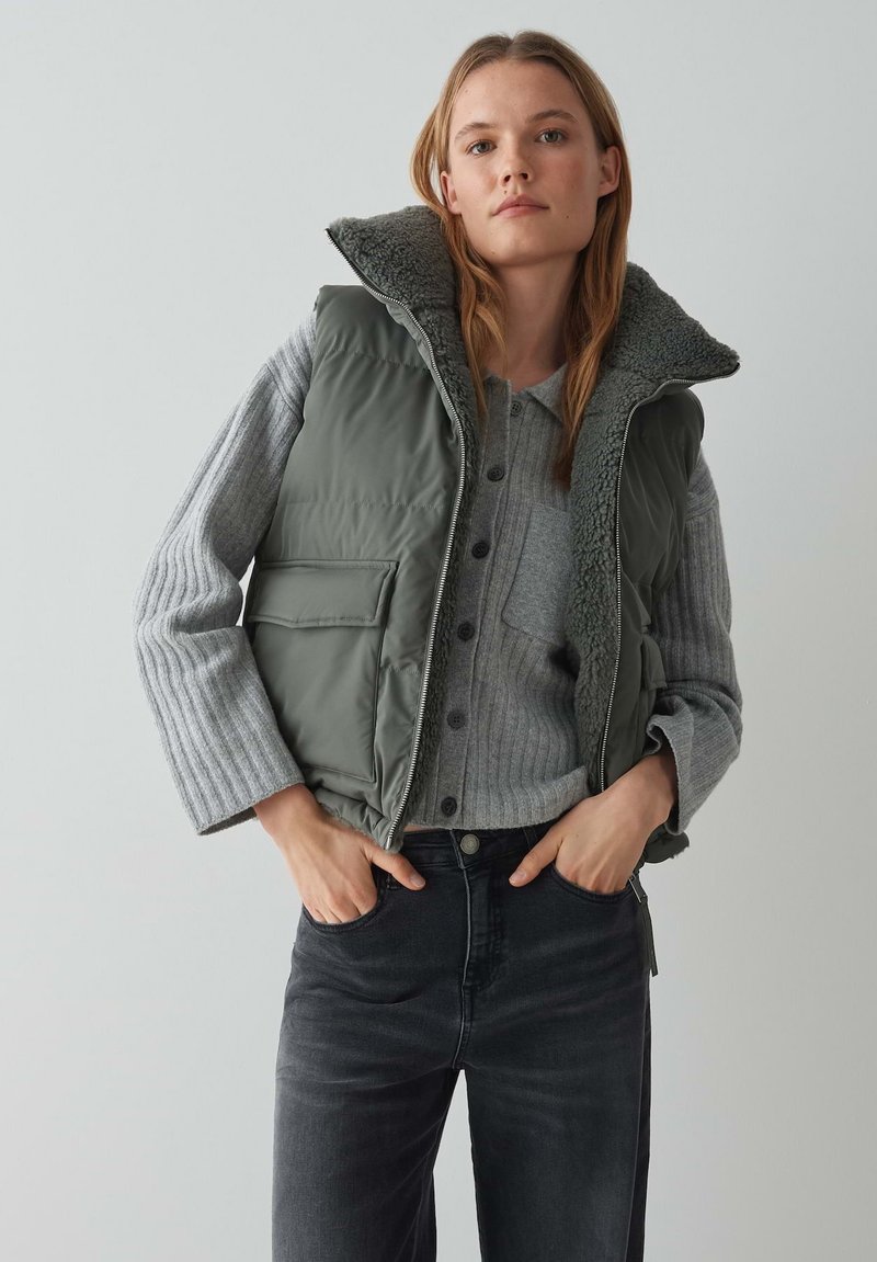 Grüne gesteppte Weste mit einem kuscheligen, fellgefütterten Kragen über einem grauen gerippten Pullover. Das Model trägt dunkle Jeans, die einen lässigen Schichtlook zeigen.