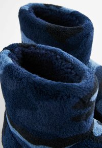 Uldne blå slippers med en blanding af mørkeblå og lyseblå nuancer. Blød tekstur, runde åbninger og et hyggeligt, plyshørt design.
