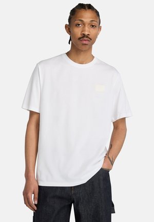 Man met gevlochten haar en snor draagt een effen wit T-shirt en donkere spijkerbroek, staand met één hand in de zak tegen een witte achtergrond.