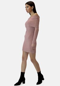 Abito a maglia a coste di colore rosa chiaro con design off-the-shoulder, maniche lunghe e silhouette attillata. Abbinato a stivali neri alla caviglia.