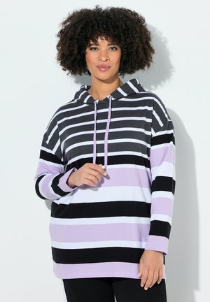 Kvinde med krøllet hår iført en sort, hvid og lavendelstribet hættetrøje med snore, stående foran en lys baggrund.