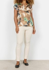 Braun-grüne Batik-Bluse mit kurzen Ärmeln, kombiniert mit beigen Slim-Fit-Hosen und schwarzen Ballerinas.