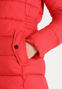 Ręka w kieszeni czerwonej pikowanej kurtki puchowej z elastycznym pasem i patką na zatrzask na przedniej kieszeni.