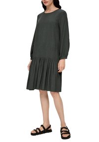 Dunkelgrünes Kleid mit rundem Ausschnitt, langen Ärmeln und einem gestuften Rüschensaum. Besitzt ein dezentes Muster und wird mit schwarzen Sandalen kombiniert.