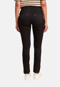 Jean noir slim à taille haute, avec cinq poches et des détails argentés, porté avec des baskets blanches sur une personne vue de dos.