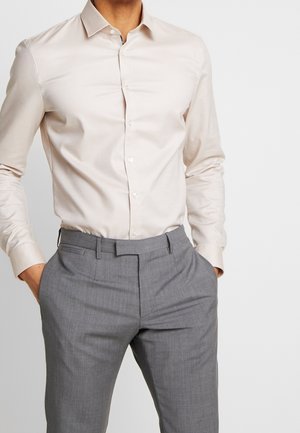 Homme portant une chemise habillée beige clair à boutons, rentrée dans un pantalon gris formel, mains dans les poches sur un fond uni.