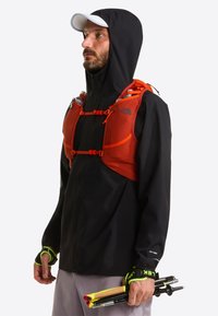 Czarna wodoodporna kurtka z kapturem i pomarańczowym szelkiem w stylu kamizelki. Model trzyma kijki trekkingowe z neonowymi akcentami.