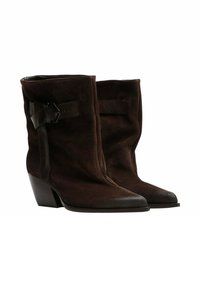 Stivaletti alla caviglia in suede marrone con un leggero tacco. Presentano una parte anteriore piatta, punta affusolata e una cinghia in pelle nera con dettaglio della fibbia.