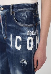 Κατεστραμμένο τζιν σε σκούρο μπλε, με λευκή επιγραφή "ICON", μικρές τρύπες και αντιθετική ραφή, σε κλασικό σχέδιο πέντε τσεπών.