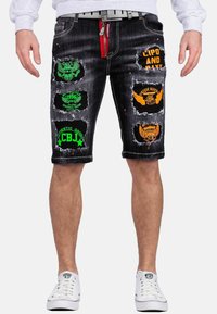 Shorts in denim neri con dettagli strappati. Presentano toppe colorate in verde, arancione e giallo. Accentazione con zip rossa sul davanti.