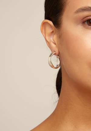 Primer plano de la oreja de una mujer que lleva un pendiente de aro grueso y plateado brillante sobre un fondo beige liso.