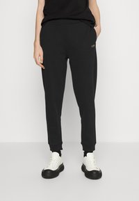 Černé tepláky s elastickým pasem a manžetami, vybavené bočními kapsami a diskrétním zlatým logem. V kombinaci s bílými teniskami.