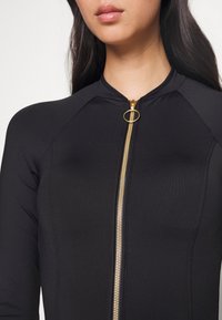 Black zip-up bodysuit with a round gold zipper pull, featuring a high neckline and long sleeves. Smooth, stretchy fabric with seam detailing.