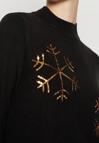 Pull noir à col rond avec un motif flocon de neige en sequins dorés. Texture douce, coupe ajustée et manches longues.