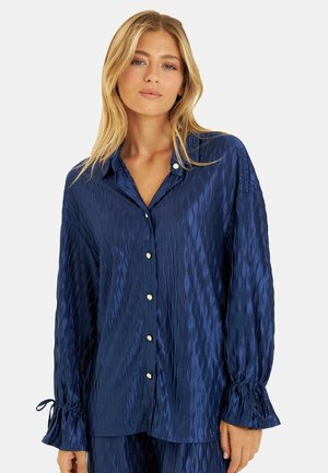 Blusa de satén azul marino con un patrón de olas texturizado, frente abotonado y puños suavemente fruncidos con lazos.