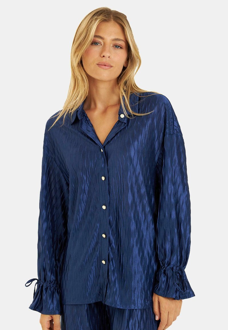 Blusa de satén azul marino con un patrón de olas texturizado, frente abotonado y puños suavemente fruncidos con lazos.