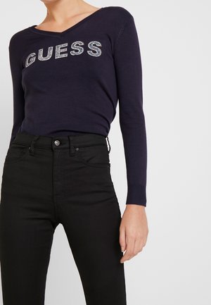 Osoba nosząca granatowy sweter z długim rękawem i dekoltem w serek z napisem „GUESS” z cyrkonii oraz czarne, wysokie jeansy.