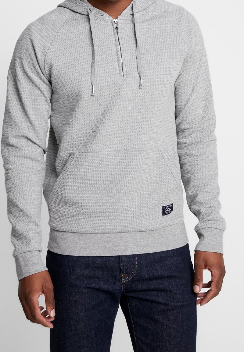 Uomo che indossa un hoodie a mezze zip grigio chiaro con tessuto a trama e tasca a marsupio, abbinato a jeans blu scuri, in piedi contro uno sfondo neutro.