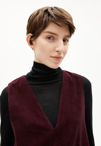 Bordeauxfarbener Cord-Westen mit V-Ausschnitt, über einem schwarzen Rollkragenpullover getragen. Die Textur ist gerippt mit einer glatten Oberfläche darunter.