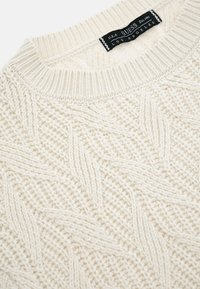 Maglione lavorato a maglia di colore avorio con motivo a trecce, scollatura rotonda e polsini a costine. Presenta un'etichetta con "GUESS" su sfondo nero.