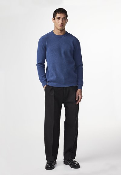 Maglione a coste blu con maniche lunghe, abbinato a pantaloni neri a gamba larga e scarpe nere lucide. Collo rotondo semplice, senza motivi.