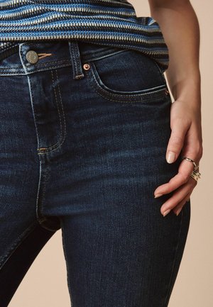 Close-up of a person wearing dark blue jeans and a striped shirt, with rings on their right hand resting on their thigh.