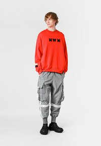 Sudadera roja con letras negras "MWM", combinada con pantalones cargo grises que tienen cremalleras y acentos blancos. Zapatos negros de textura tipo slip-on.