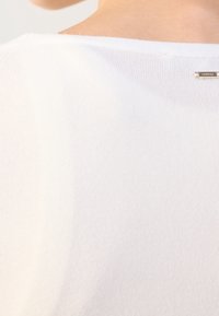 Gros plan sur l'épaule et le haut du dos d'une personne portant un haut blanc côtelé, épaules dénudées, avec une petite étiquette métallique de marque.