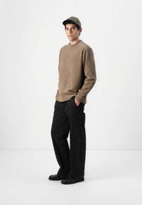Maglietta marrone a righe con maniche lunghe, pantaloni neri a gamba larga e berretto a quadri. Footwear neri completano l'outfit. Sfondo bianco semplice.