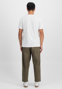 Bílá bavlněná tričko s klasickým střihem, kombinované s olivově zelenými kalhotami. Model stojí zády, což odhaluje jednoduchý a čistý design.