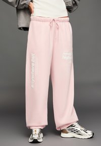 Ružové tepláky s elastickým pásom a manžetami na členkoch, so bielymi nápismi po bokoch. Nosené so striebornými a bielymi teniskami.