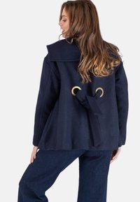 Marineblauwe wolmix jas met oversized kraag, achterkant met gouden ringen en een stoffen strik, gecombineerd met donkere spijkerbroek.