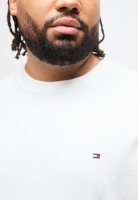 Άνδρας με γένια και κότσους, που φοράει λευκό πουκάμισο με μικρό κόκκινο, λευκό και μπλε ορθογώνιο λογότυπο στο στήθος.