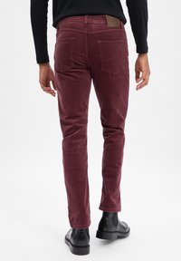 Mann in schmal geschnittenen burgunderfarbenen Cordhosen und schwarzen Leder Chelsea Boots, vor einem schlichten weißen Hintergrund stehend.