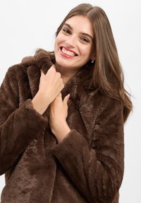 Bruine nepbontjas met hoge kraag, textuur op de oppervlakte en lange mouwen. Het model houdt de kraag dicht bij haar gezicht.