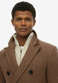 Abrigo de espiga marrón con diseño de doble botonadura y botones negros, superpuesto a un suéter de punto trenzado color crema.