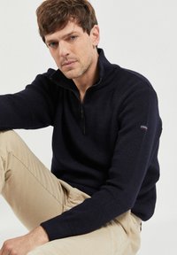 Granatowy sweter z zamkiem błyskawicznym w strukturze dzianiny. Posiada długie rękawy oraz zamek z przodu. Wykończony logotypem marki na rękawie.