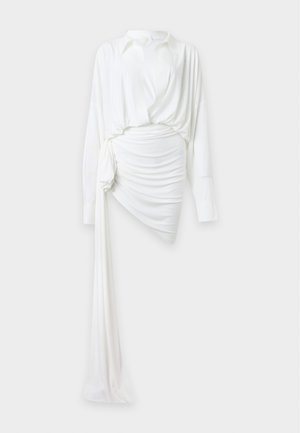 Femme portant un chemisier blanc à manches longues avec un décolleté plongeant en V et une jupe blanche asymétrique froncée, avec des sandales noires à brides sur fond blanc.