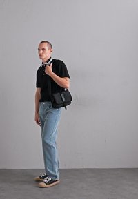 Bărbat îmbrăcat cu un tricou polo negru, blugi de culoare albastră deschisă, adidași negri, purtând o geantă de umăr Moschino neagră, stând în picioare lângă un perete gri simplu.