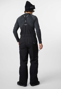 Black waterproof snow overalls with adjustable straps, zippered pockets, and elastic waistband, worn over a dark gray long-sleeve shirt.