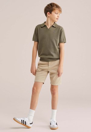 Fant, ki nosi olivno zeleno polo majico s kratkimi rokavi, bež kratke hlače, bele nogavice in bele superge s črnimi črtami, stoji in gleda desno.