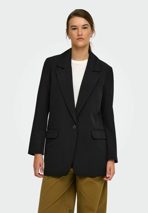 Vrouw draagt een zwarte blazer met één knoop over een wit overhemd en mosterdkleurige broek, staand tegen een effen lichte achtergrond.