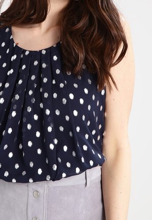 Femme portant un chemisier sans manches bleu marine à pois argentés, rentré dans une jupe boutonnée gris clair, cheveux bruns ondulés mi-longs visibles.