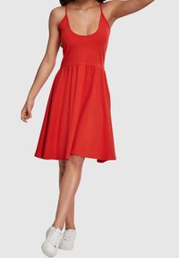 Robe rouge sans manches avec un décolleté rond, un corsage ajusté et une jupe évasée. Associée à des baskets blanches. Fabriquée en tissu doux et extensible.