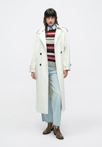 Lang hvid trenchcoat med sorte knapper, båret over en flerfarvet stribet sweater, sat sammen med lyseblå vidde jeans og brune ankelstøvler.