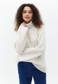 Cremfarbener Rollkragenpullover mit einer weichen, gerippten Textur, weit geschnittenen Ärmeln und einem asymmetrischen Saum. Kombiniert mit dunkelblauen Cordhosen.