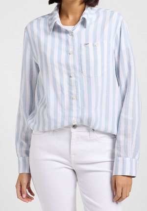 Vrouw draagt een lichtblauw-wit verticaal gestreept blouse met witte broek, blouse gedeeltelijk ingestopt.