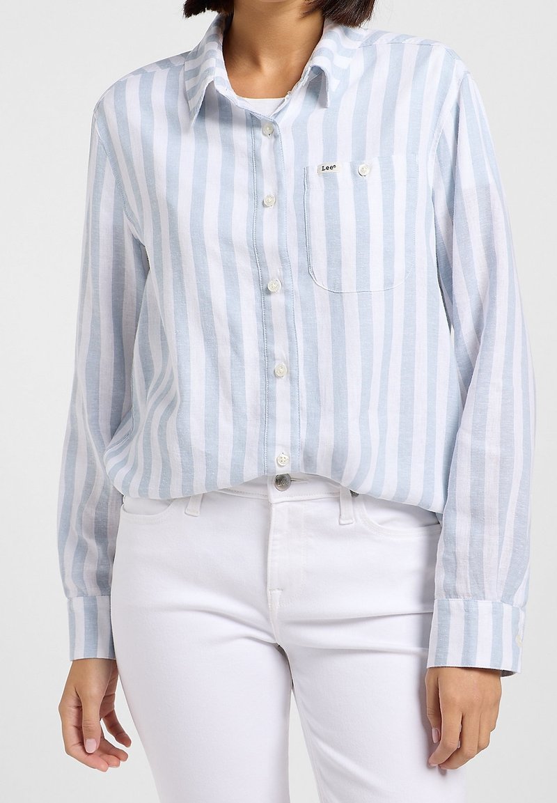 Femme portant une chemise boutonnée à rayures verticales bleu clair et blanc, avec un pantalon blanc, la chemise partiellement rentrée.