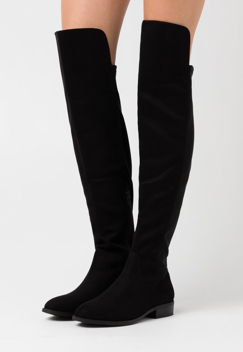 Botas negras sobre la rodilla con una textura suave, similar al ante. Cuentan con una cremallera lateral y un tacón bajo y ancho. Diseño de punta redondeada y suave.