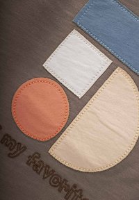 Tessuto di cotone in marrone tenue presenta applicazioni colorate: cerchio arancione, quadrato bianco e forma arrotondata in beige con accento blu.